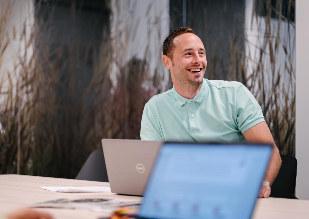 Medewerker kijkt lachend en zit aan een tafel met 2 laptops