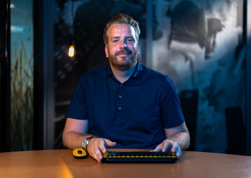 mannelijke medewerker kijkt vriendelijk in de camera met toetsenbureau op zijn bureau