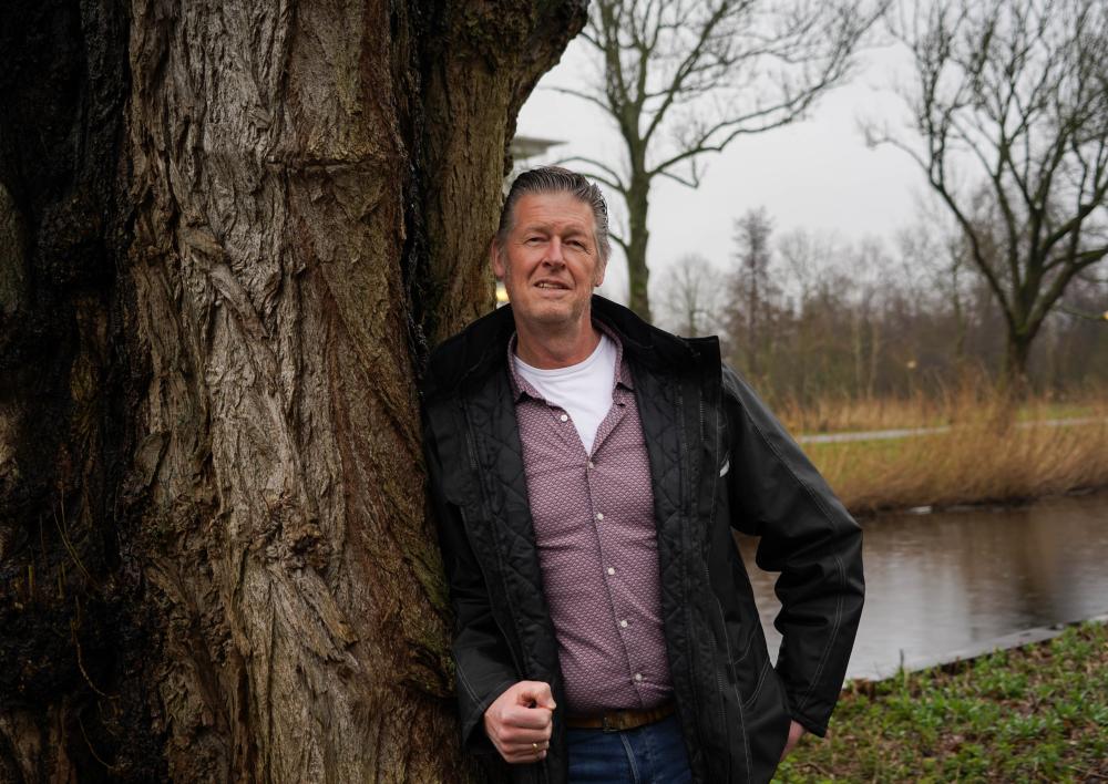 Medewerker leunt tegen een grote boom met een slootje op de achtergrond