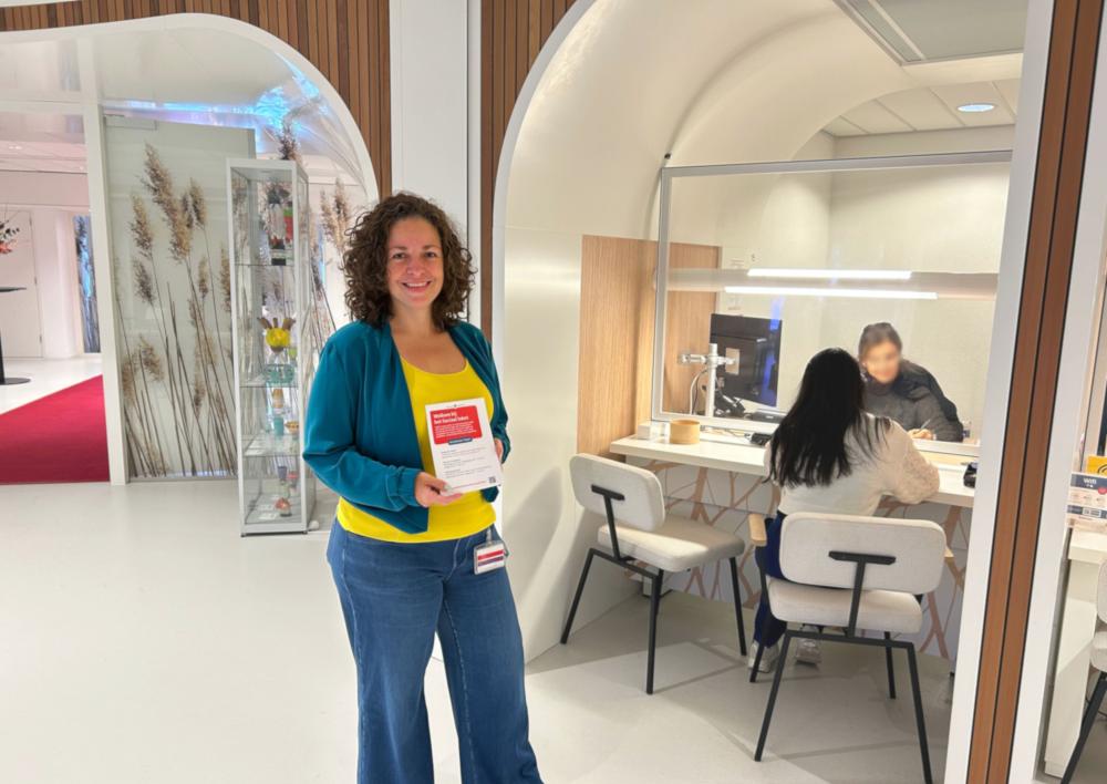 Maaike met folder van sociaal loket voor het loket. Bij het loket spreekt een medewerker met een bezoeker