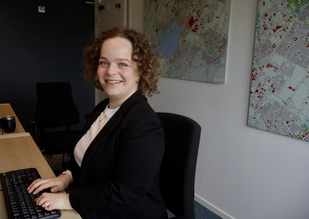Maxime achter de computer met plattegronden op de muur als achtergrond