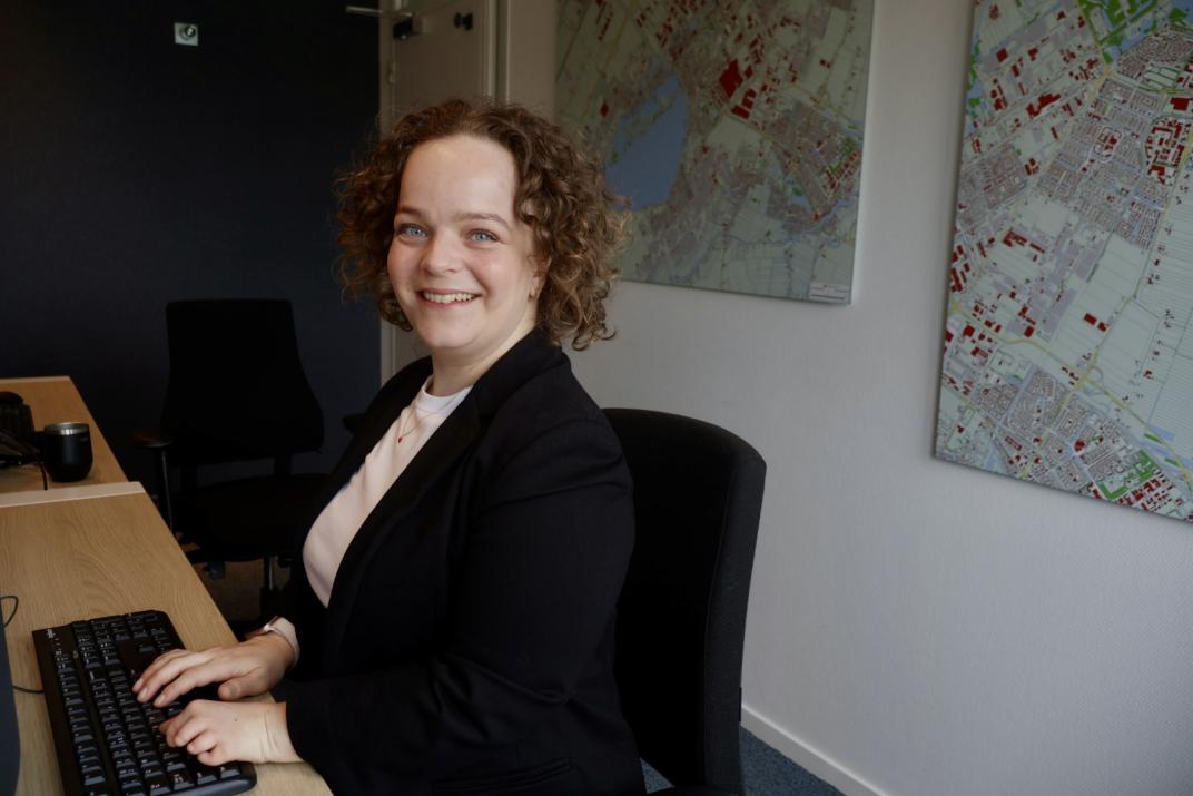 Maxime achter de computer met plattegronden op de muur als achtergrond