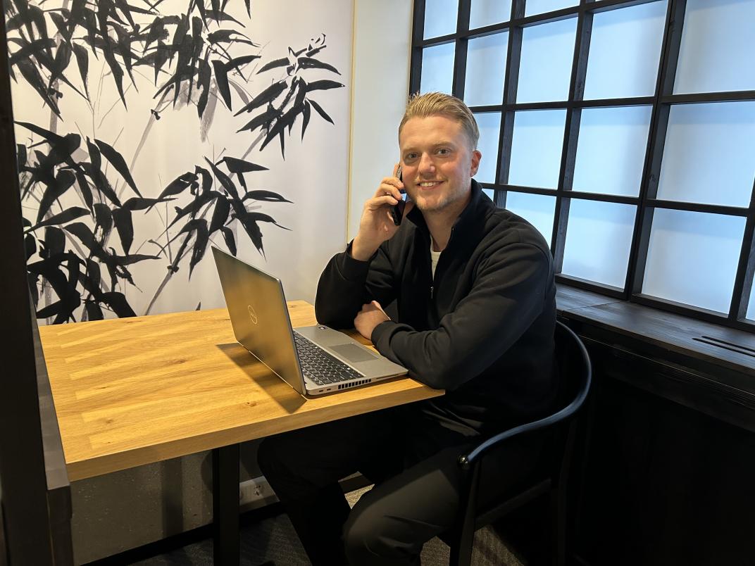 Ruben zit met laptop aan tafel, belt en kijkt in de camera
